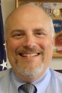 A medium skin toned man who is bald with a grey beard. He is wearing a blue shirt and grey tie