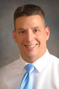 A medium light skin toned man with brown hair in a buzzcut. He is wearing a white shirt and blue tie