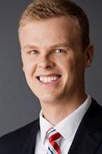 A light skin toned man with short blond hair wearing a white shirt, red tie and black suit jacket