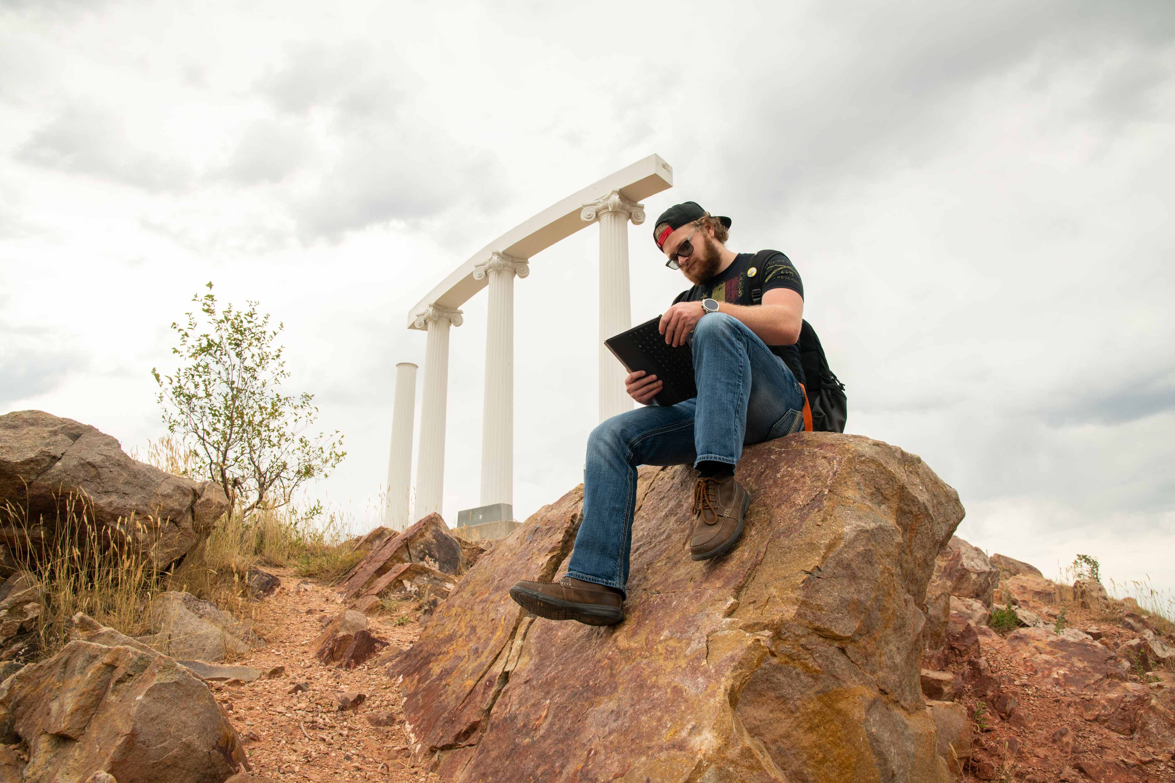 Student using a laptop sitting on a rock with the Red Hill pillars in the background.