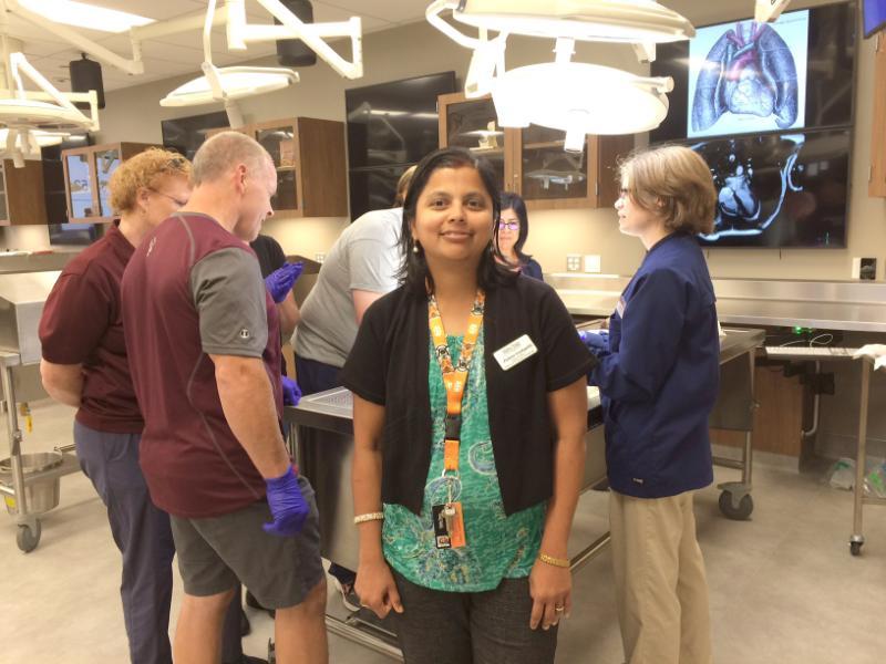 Doctor stands in front of anatomy table with students working in background.