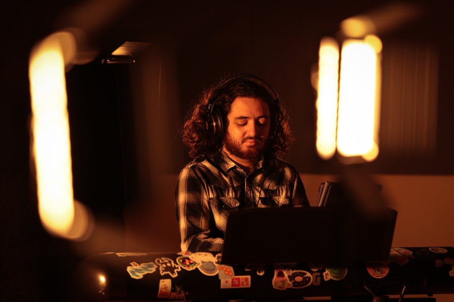 Gabe Lowman wears headphones. He sits at a keyboard playing the keyboard in a recording studio with warm lighting.