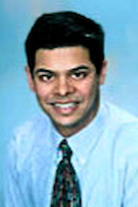 A medium-dark skin toned man with black hair. He is wearing a light blue shirt and a black tie. He is smiling