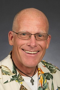 A medium skin toned man who is bald. He is wearing glasses, a cord with a cross on it, a white undershirt and a beige Hawaiian patterned button down.