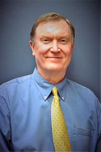 A light skin toned man with blond hair. He is wearing a blue button down and a yellow tie.