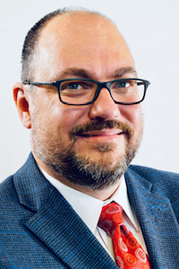 A man with medium skin tone and balding brown hair. He has a brown beard and mustache. He is wearing black glasses, a white button down, a red tie and a dark blue suit jacket.