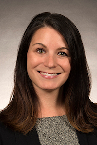 A woman with medium skin tone and brown hair. She is wearing a grey shirt and a black jacket.