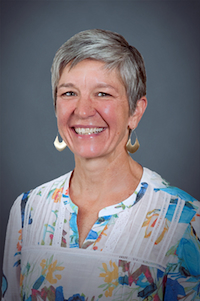 A medium skin toned woman with grey short hair. She is wearing gold earrings and a patterned blouse. She is smiling.