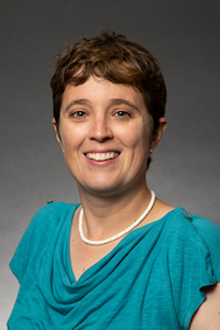 A woman with medium skin tone and short brown hair. She is wear a white necklace and a teal shirt. She is smiling