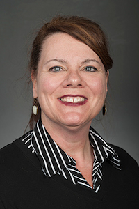 A woman with light skin tone and reddish-brown hair. She is wearing earrings and a white and black striped collared shirt with a black sweater over it. She is smiling.