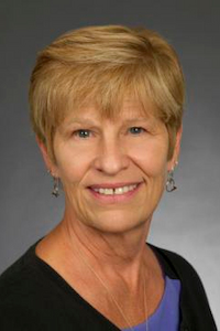 A medium skin toned woman with short blond hair. She is wearing earrings, a necklace, a purple shirt and a black cardigan. She is smiling