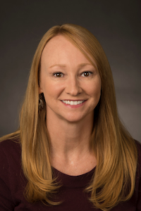 A medium skin toned woman with blond hair. She is wearing earrings and a purple shirt. She is smiling