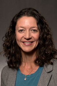 A woman with medium skin tone and brown hair. She is wearing a necklace, blue shirt, and grey blazer. She is smiling