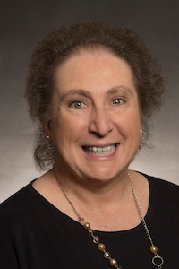 A medium skin toned woman with grey curly hair. She wearing earrings, a necklace, and a black shirt.