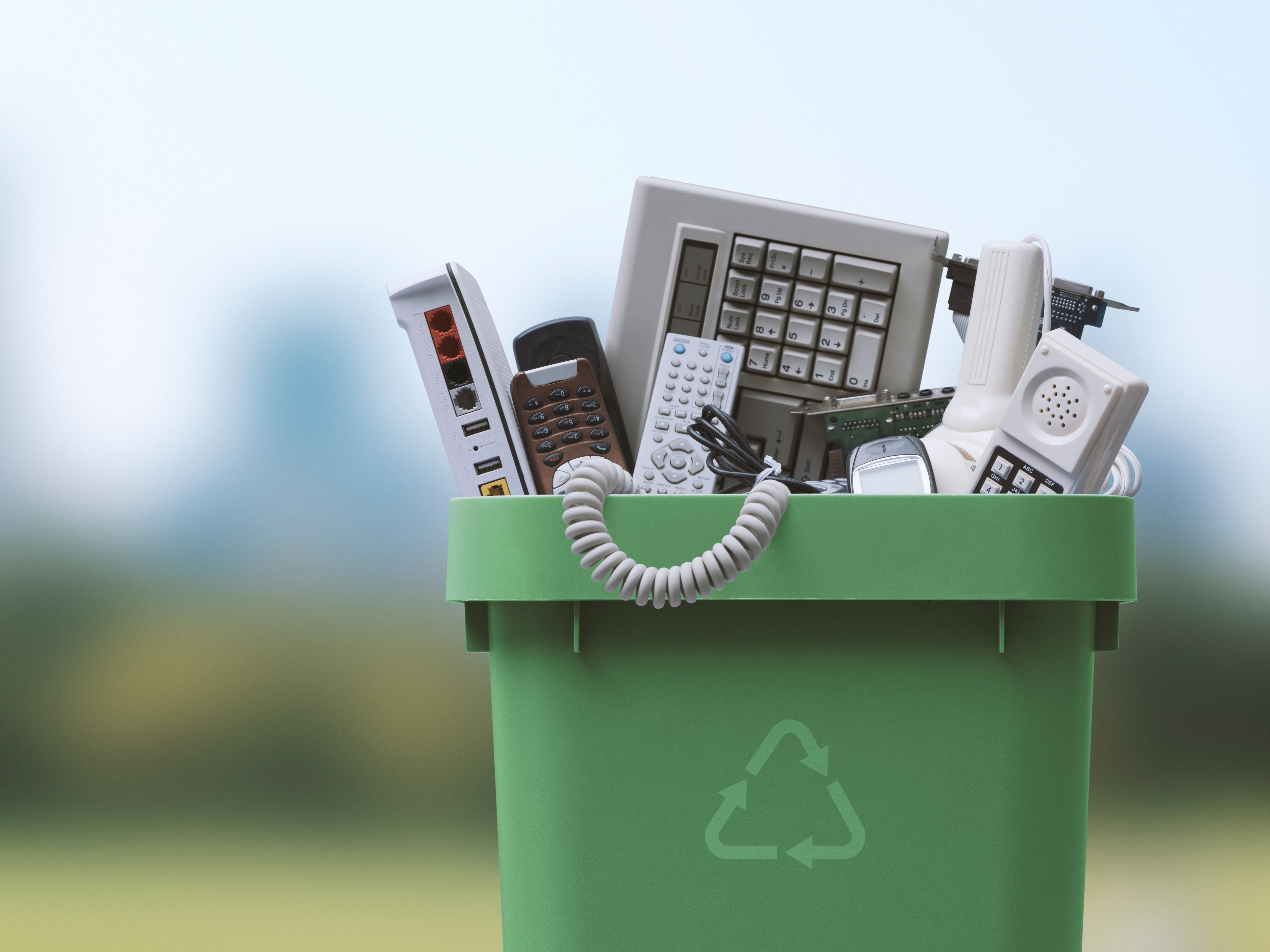 green recycling bin full of discarded electronics against a blurred outdoor background