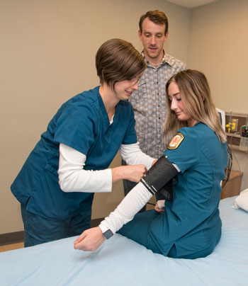 Two nursing students learn how to take blood pressure while an instructor observes in the background.
