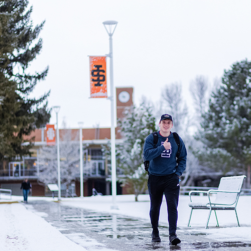 A student walking near the SUB in Winter, gesturing thumbs up.