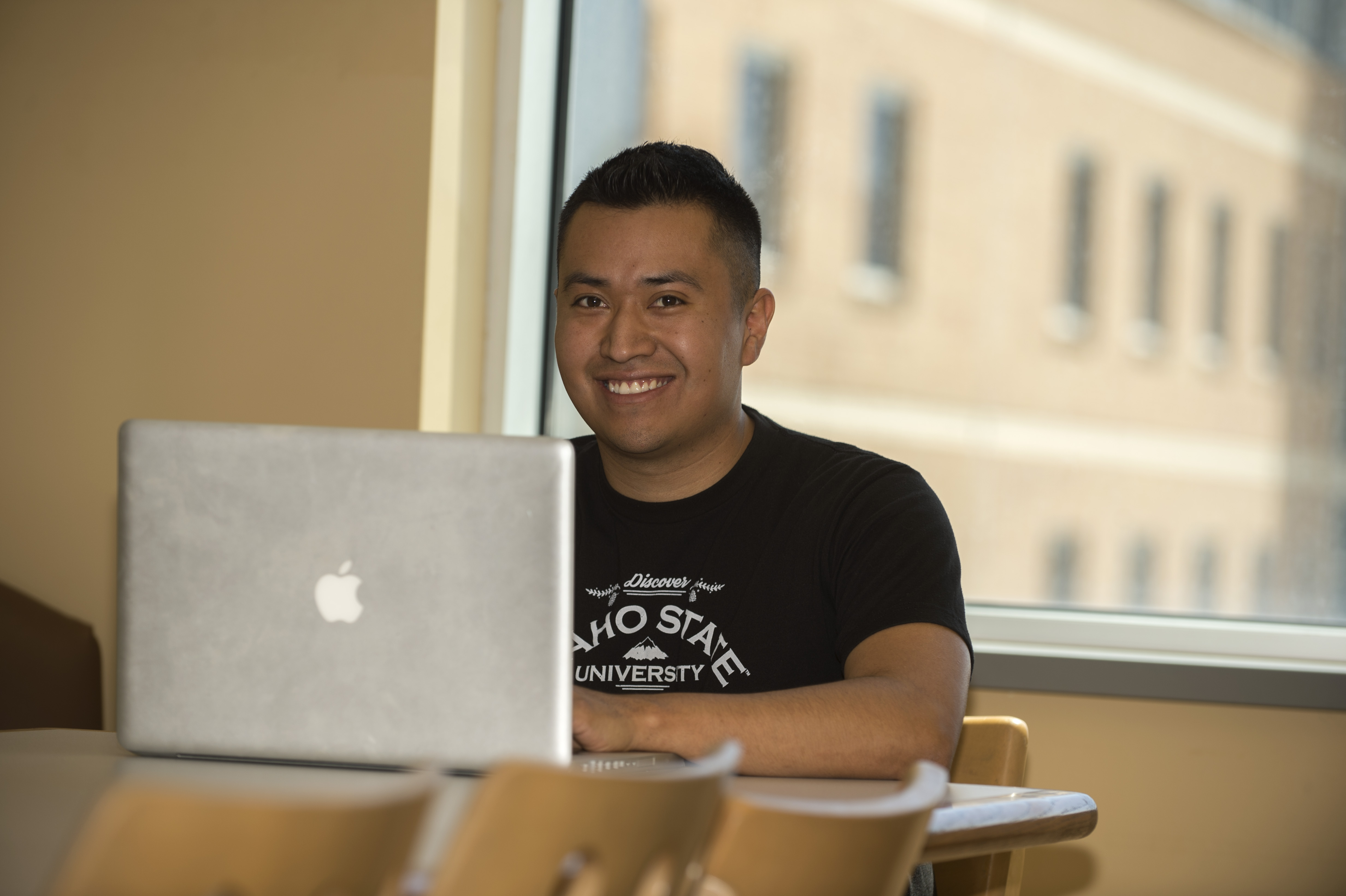 A student sitting indoors in front of a laptop, smiling at the camera.