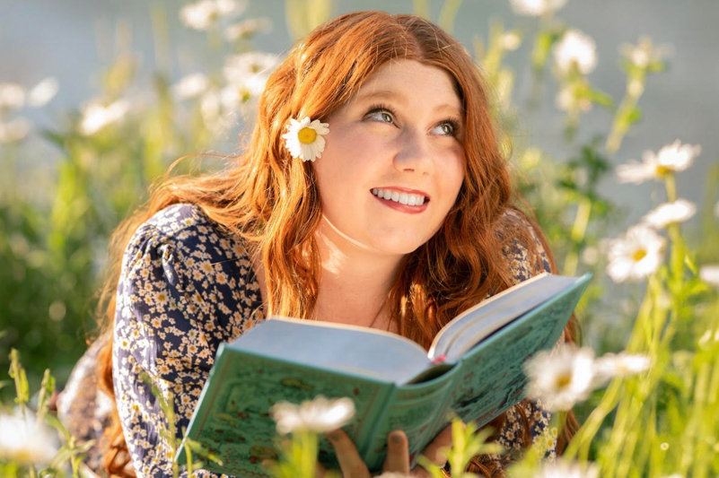 A close-up, dreamlike photo of a woman with long, wavy red hair lying in a field of white daisies. She is looking upward with a joyful smile, and a single daisy is tucked behind her ear. She holds an open, light-blue book in front of her. The scene is bathed in warm, soft sunlight, with many flowers blurred in the foreground and background.