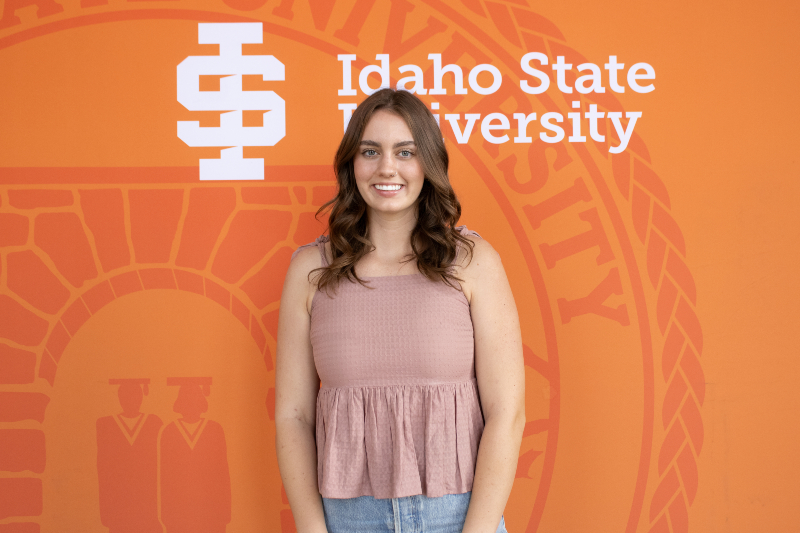 A medium shot of a young woman with wavy brown hair and a bright smile, standing in front of an orange background. She is wearing a sleeveless, dusty pink peplum top and blue jeans. Behind her is a large Idaho State University logo, featuring the 
