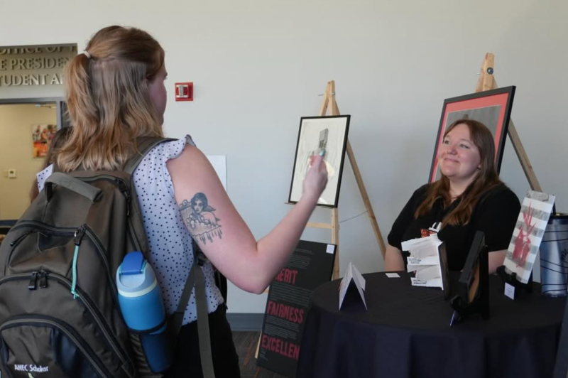 Two young women interact at an art display table indoors. One woman stands in the foreground with her back to the camera, wearing a white polka-dot sleeveless top, a large olive green backpack with a blue water bottle, and a distinct tattoo on her upper arm. She is gesturing toward the second woman, who is seated behind a black-clothed table and smiling warmly. On the table and surrounding easels