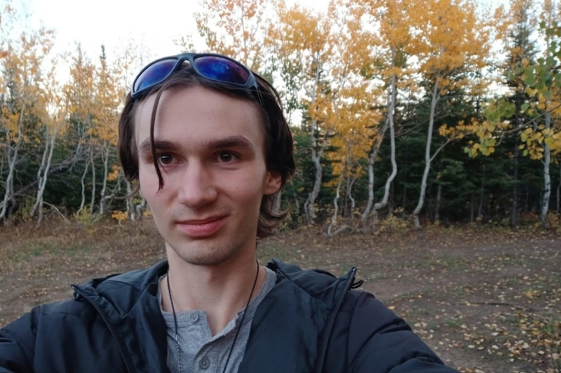 A young man with brown hair and a slight smile takes a selfie in a wooded area. He is wearing a grey henley shirt, a black jacket, and a beaded necklace, with dark sunglasses resting on top of his head. The background features several trees with bright yellow autumn leaves under a pale sky.