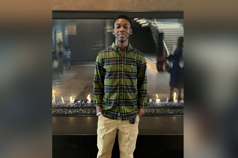 A young man with short black hair stands with his hands in his pockets, smiling slightly. He is wearing a green, black, and white horizontal-striped button-down shirt and tan cargo pants. He is posed in front of a wide, modern indoor fireplace with small blue and yellow flames. The background shows a dimly lit lobby with a staircase and a polished floor.