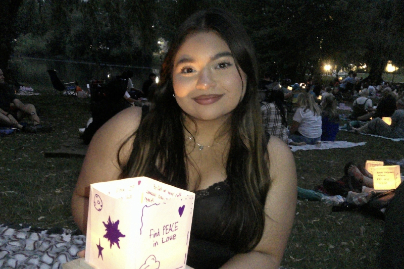 A young woman with long, dark hair and a gentle smile poses at an evening event. She is holding a glowing paper lantern decorated with the handwritten message 