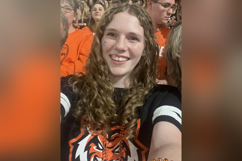 A young woman with long, curly light brown hair smiles enthusiastically in a crowded stadium setting. She is wearing a black t-shirt with orange and white stripes on the sleeves and a tiger logo on the front, accented by orange beaded necklaces. She has a small tiger mascot sticker on her cheek. The background is filled with other fans wearing orange, creating a vibrant game-day atmosphere.