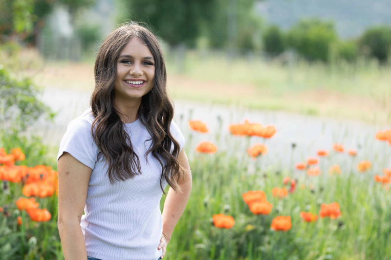 A young woman with long, wavy dark brown hair and a bright smile stands in a field. She is wearing a light grey or lavender ribbed t-shirt and has one hand on her hip in a relaxed pose. She is surrounded by tall green grass and vibrant, blooming orange poppies, with a soft-focus background of trees and a distant path under bright, natural light.