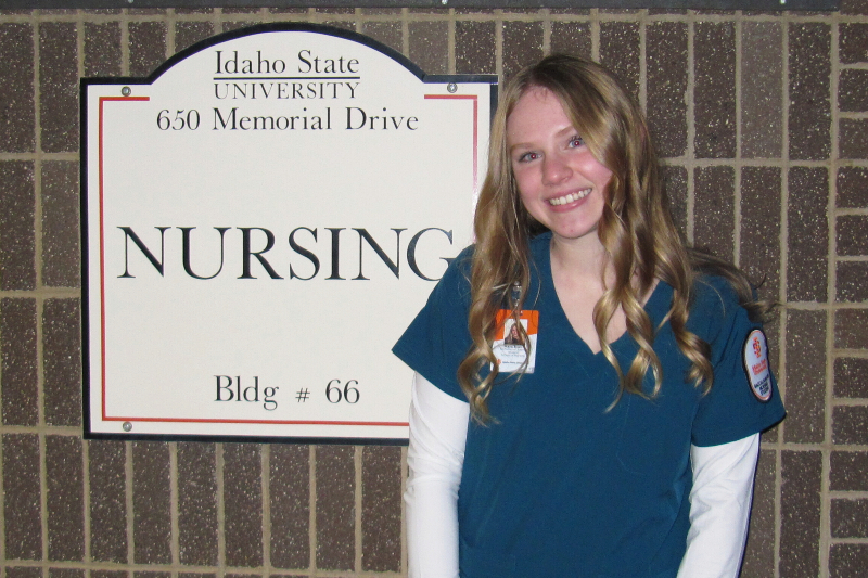 A young woman with long, wavy blonde hair and a bright smile stands in front of a brick wall next to a sign. She is wearing dark teal nursing scrubs over a white long-sleeved shirt, complete with an identification badge and a university patch on her sleeve. The sign beside her reads 