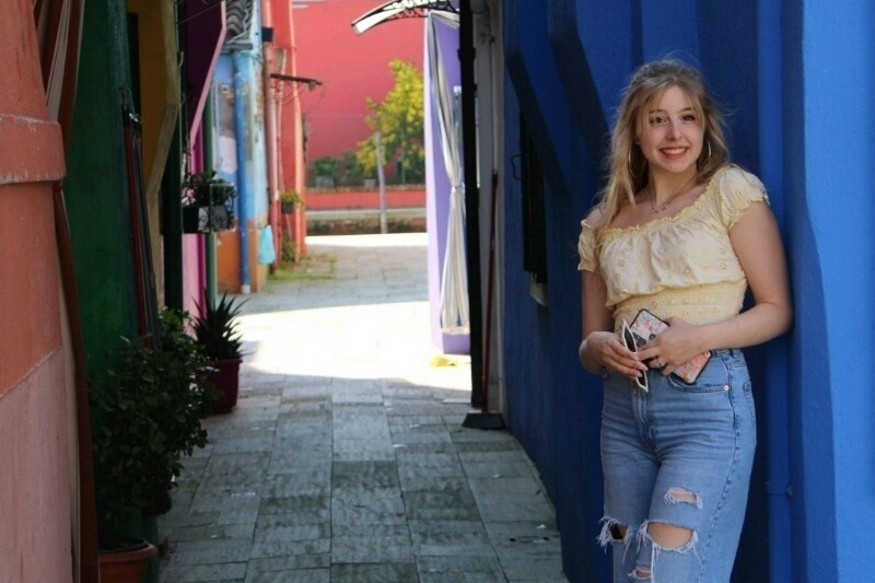 A young woman with blonde hair and a joyful smile leans against a bright blue wall in a narrow, colorful alleyway. She is wearing an off-the-shoulder light yellow top and ripped blue jeans, holding a phone and sunglasses in her hands. The background features a stone-paved path stretching past buildings painted in vibrant shades of pink, green, and red.