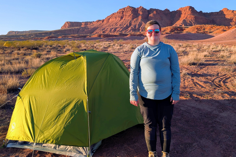 A young woman stands beside a bright lime-green tent in a desert landscape. She is wearing a light blue long-sleeved hooded sun shirt, black hiking pants, and red-framed mirrored sunglasses. The background features expansive dry brush and dramatic red rock mesas glowing under the warm light of a setting or rising sun.