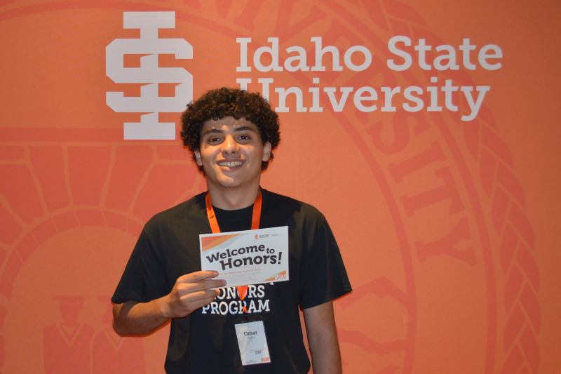 A young man with dark, curly hair and a wide, happy smile stands in front of a bright orange Idaho State University backdrop. He is wearing a black t-shirt and an orange lanyard, and he is holding up a card that says 