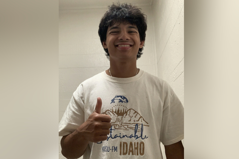 A young man with messy dark hair and a wide, friendly smile gives a 