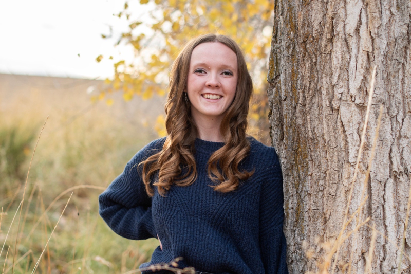 A young woman with long, wavy brown hair and a bright smile leans against a large, textured tree trunk. She is wearing a navy blue oversized ribbed sweater and one hand is resting on her hip. The background is a soft-focus autumn landscape with golden field grass and yellow leaves illuminated by warm, natural light.