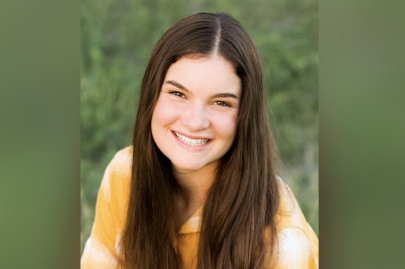 A close-up portrait of a young woman with long, straight dark brown hair and a bright, warm smile. She is wearing a yellow and white plaid button-down shirt. The background is a soft-focus field of green grass, with natural light highlighting her features.
