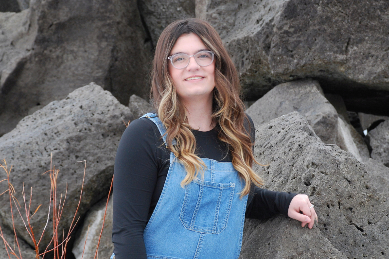 A young woman with long, wavy brown hair and blonde highlights smiles while posing outdoors. She is wearing clear-framed glasses, a black long-sleeved shirt, and blue denim overalls. She leans against a background of large, jagged grey basalt rocks.