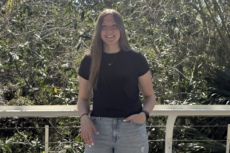 A young woman with long, straight brown hair and a friendly smile stands outdoors. She is wearing a black t-shirt, light-wash blue jeans, and a delicate silver necklace. She has one hand in her pocket and leans against a white metal railing, with a dense background of sunlit green trees and foliage.
