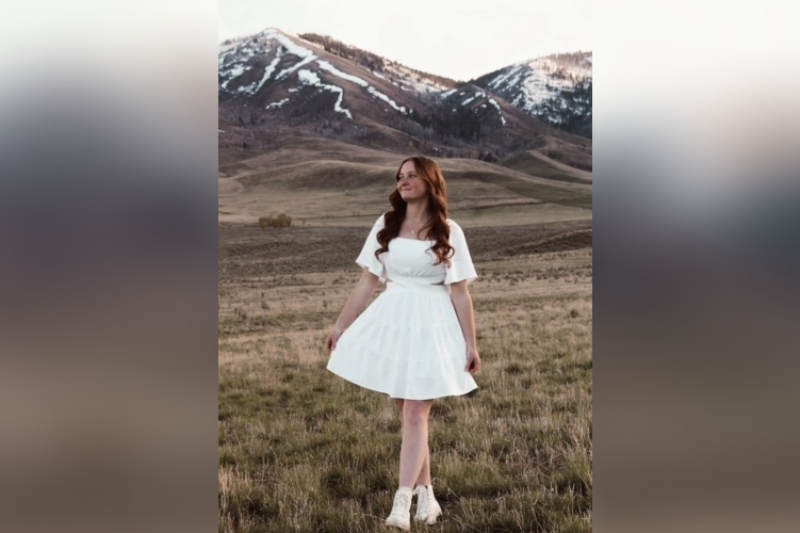 A young woman with long, wavy auburn hair smiles as she looks off to the side, posing in a vast field. She is wearing a short, tiered white dress with flutter sleeves and cut-out details at the waist, paired with off-white high-top sneakers. The background features a dramatic mountain range with patches of snow under an overcast sky.