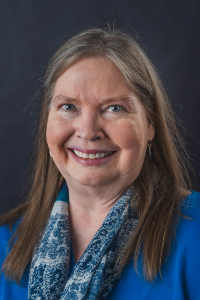 A close-up portrait of a mature woman with light brown and grey hair and blue eyes smiling warmly. She is wearing a bright blue top paired with a blue and white patterned scarf. The photo is taken against a solid dark grey studio backdrop with professional, even lighting.