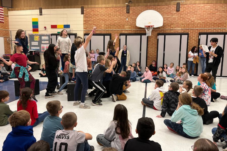 Elementary students sit in a circle in the school gym watching theatre students perform
