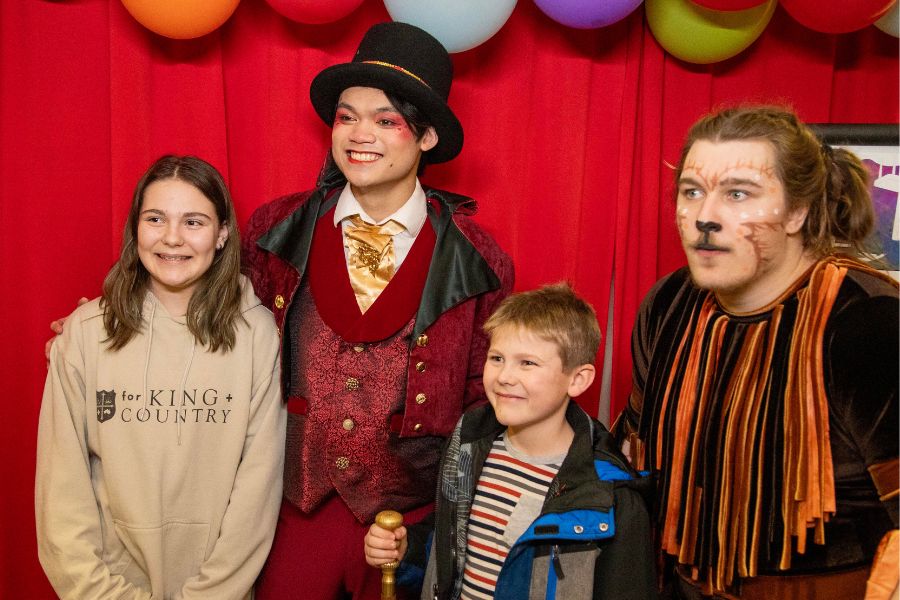 2 cast members in costume stand with 2 children from the community in front of a red backdrop and colorful balloons