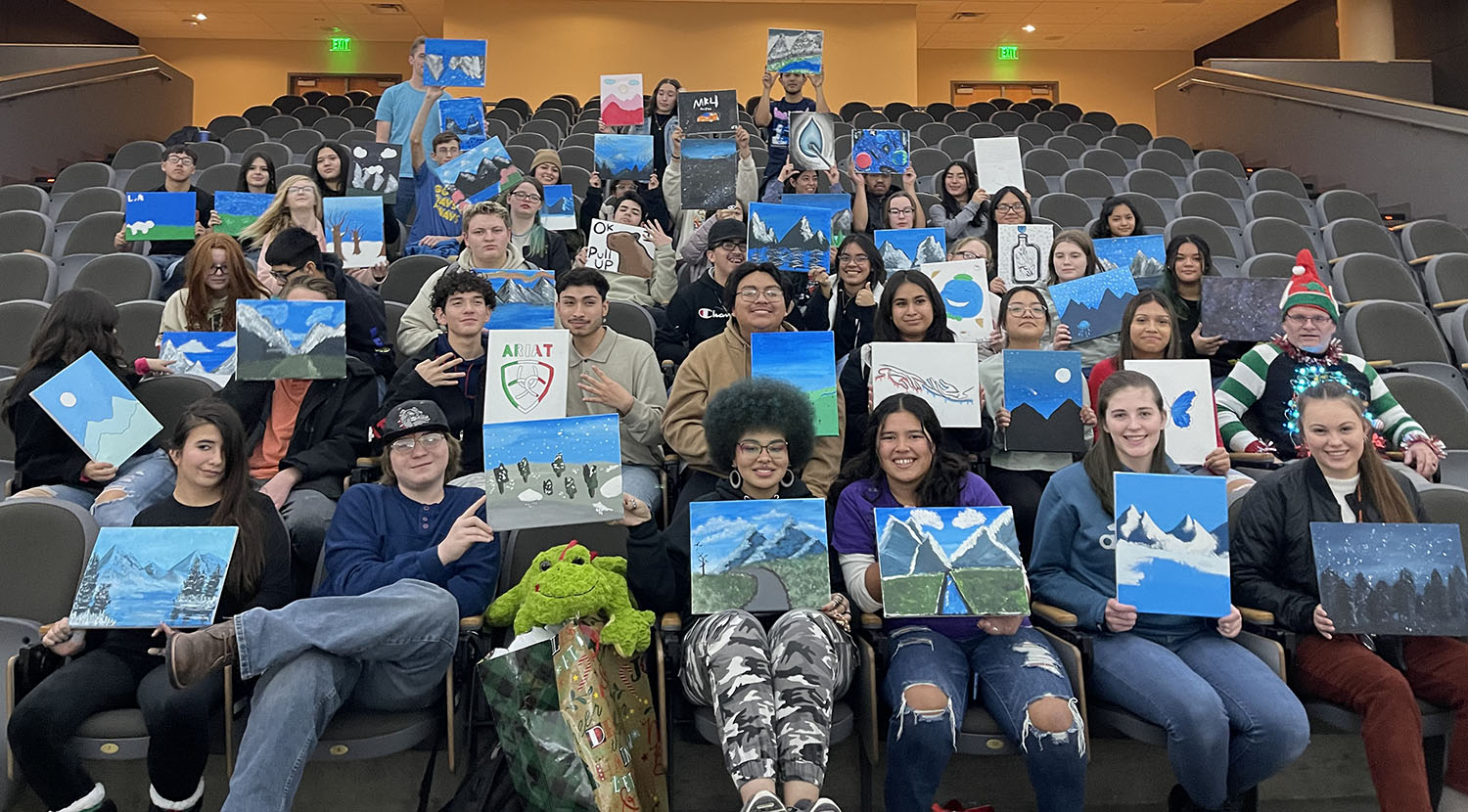 Upward Bound Programs students in an auditorium showing the paintings that they made during a Saturday Academy session. Most of the paintings are of mountains in winter.