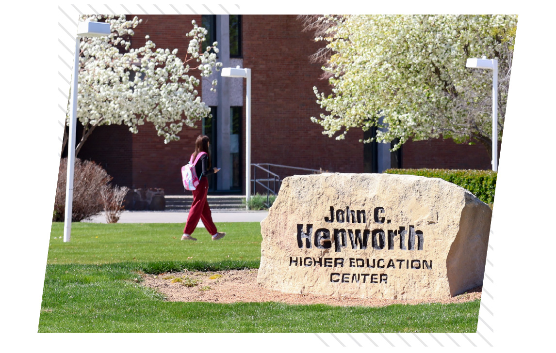 A student wearing a backpack walks across the lawn near the John C. Hepworth Higher Education Center on the College of Southern Idaho campus in Twin Falls. Trees in bloom and campus landscaping surround the brick academic building in the background.
