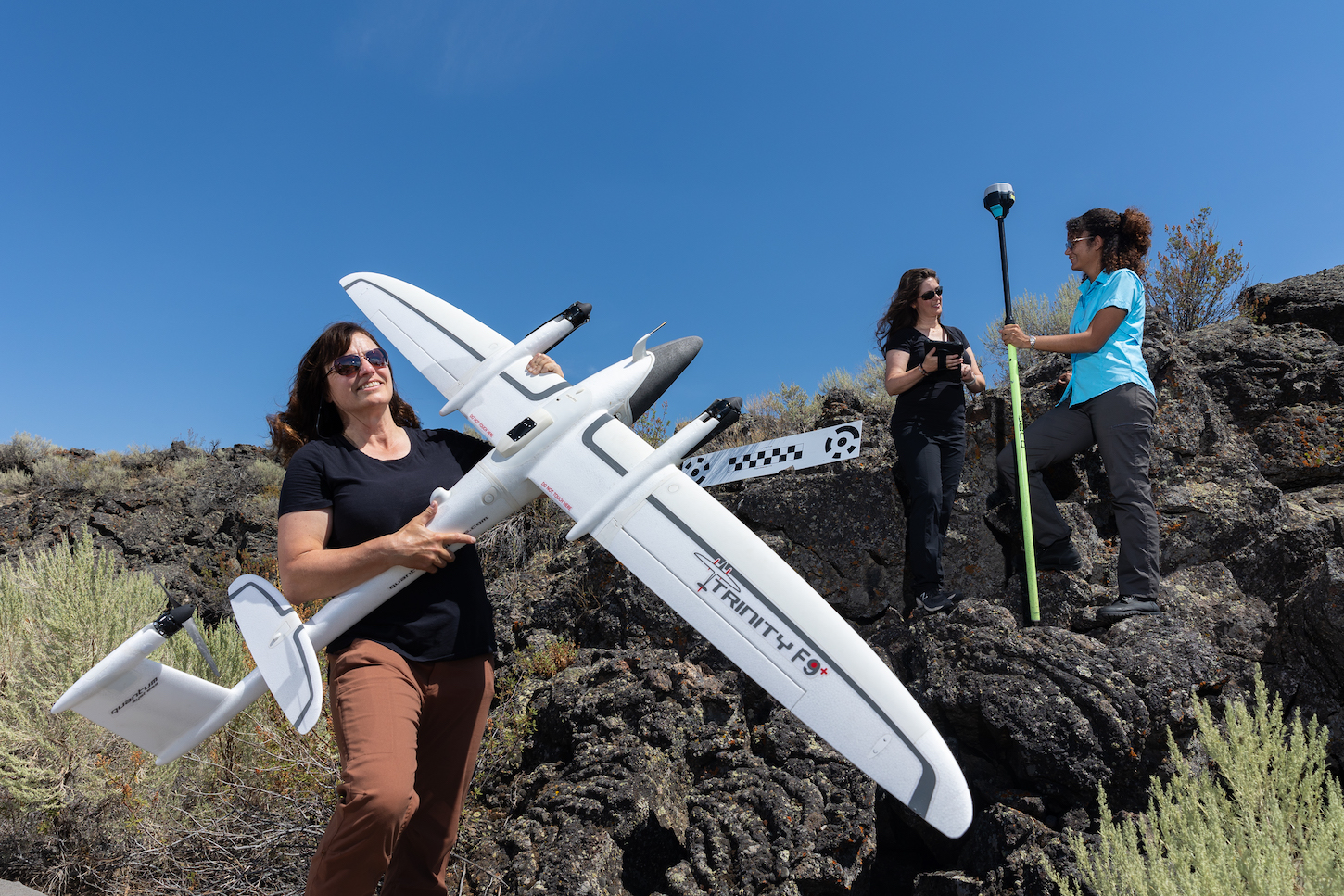 three women stand on a rocky area, one with a small plane.