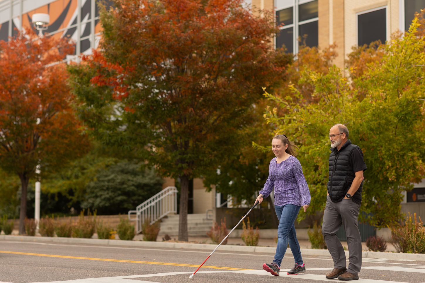 A blind woman walks across the street with a man