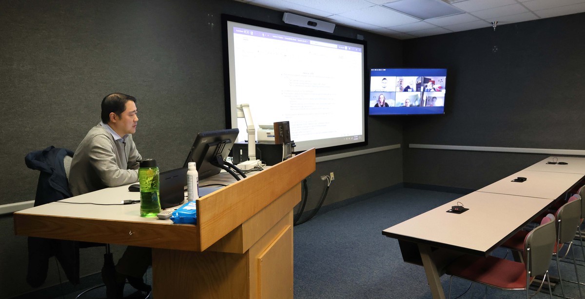 a man sits at a computer while teaching a group of students on another screen