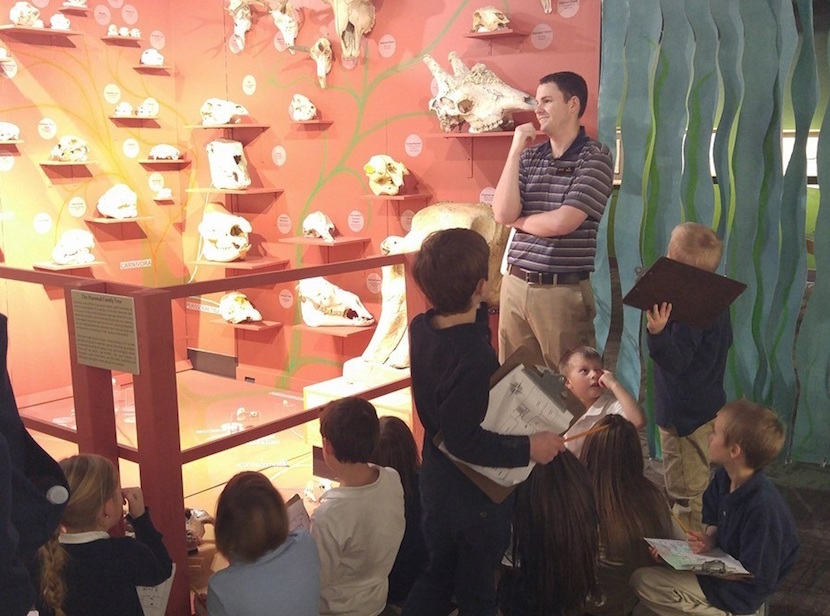 Logan Brigg standing in front of an Idaho Museum of Natural History display of various animal skulls with a group of children sitting in the foreground. 