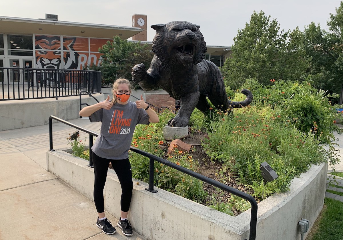 Emma Watts with mask giving two thumbs up by the Bengal sculpture on the Quad.